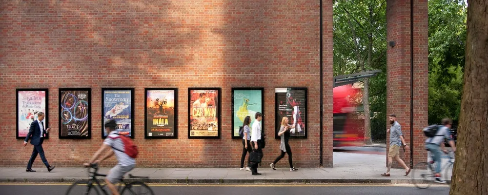 A photo of the outside wall of Sadler's Wells Theatre building with show posters and pedestrians and a cyclist passing by