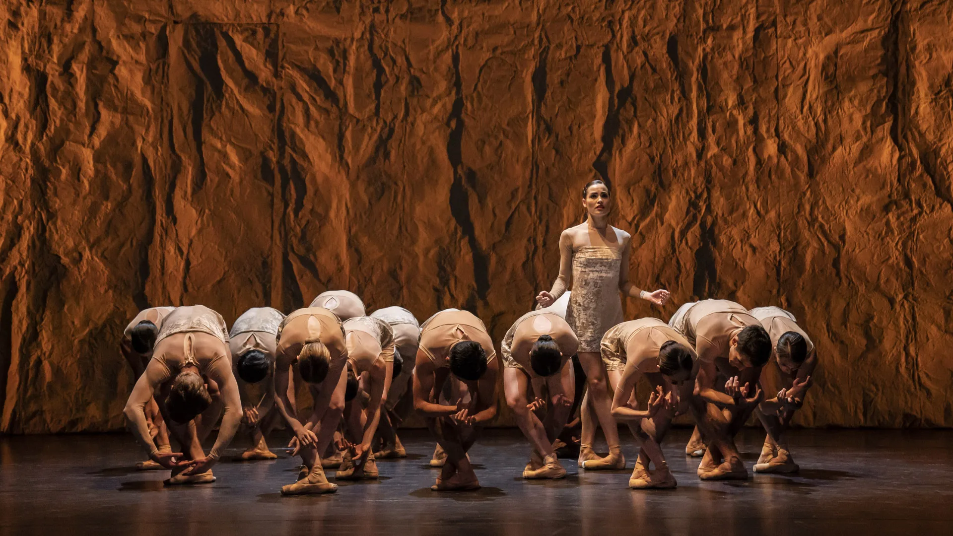 A group of ballerinas all hunched over facing the floor. One solo female dancer stands and looks into the audience