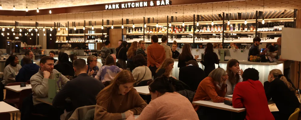 People sit at tables and interact in a busy restaurant with a well-lit bar area labeled 