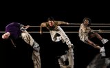 A group of five dancers lean dramatically against thick ropes on a glossy black stage, captured in dynamic, off‑balance poses.