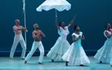 A group of dancers walk across the stage wearing white, holding white props high in the air
