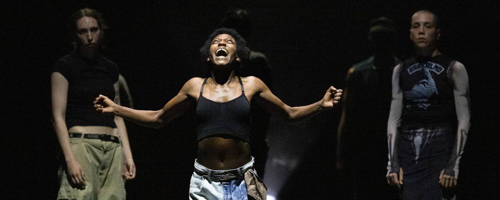 Dancer extending their arms during an expressive performance with other performers in the background.