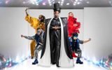 A group of four people in colorful, futuristic outfits pose energetically against a bright, sparkly background.