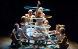 A group of performers gather around a central dancer, who is surrounded by glowing blue and yellow hoops. They wear colourful costumes and pose on a dark stage, creating a playful and striking scene.