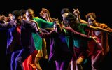 A group of contemporary dancers in brightly coloured clothes move together against a black background, with their arms reaching out in different directions. Their energetic poses create a strong sense of movement and unity.