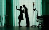 Two people stand facing each other in silhouette in a softly lit room, surrounded by hospital beds and medical equipment. Green curtains fill the background, creating a dreamlike atmosphere.