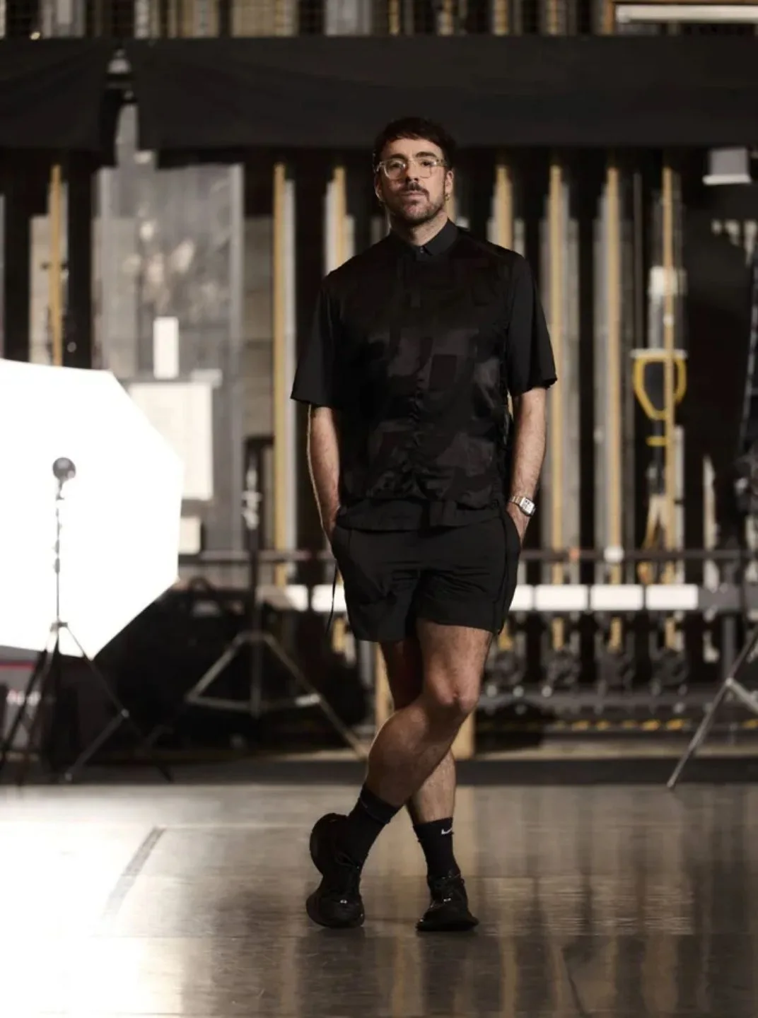 Person in an all black outfit standing on a studio floor, with stage rigging and lighting equipment in the background.